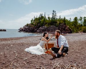 A couple shares champagne during beach picnic near rocky cliffs on a sunny day.