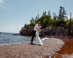 A romantic embrace on a scenic beach with rocky cliffs and evergreen forest backdrop.