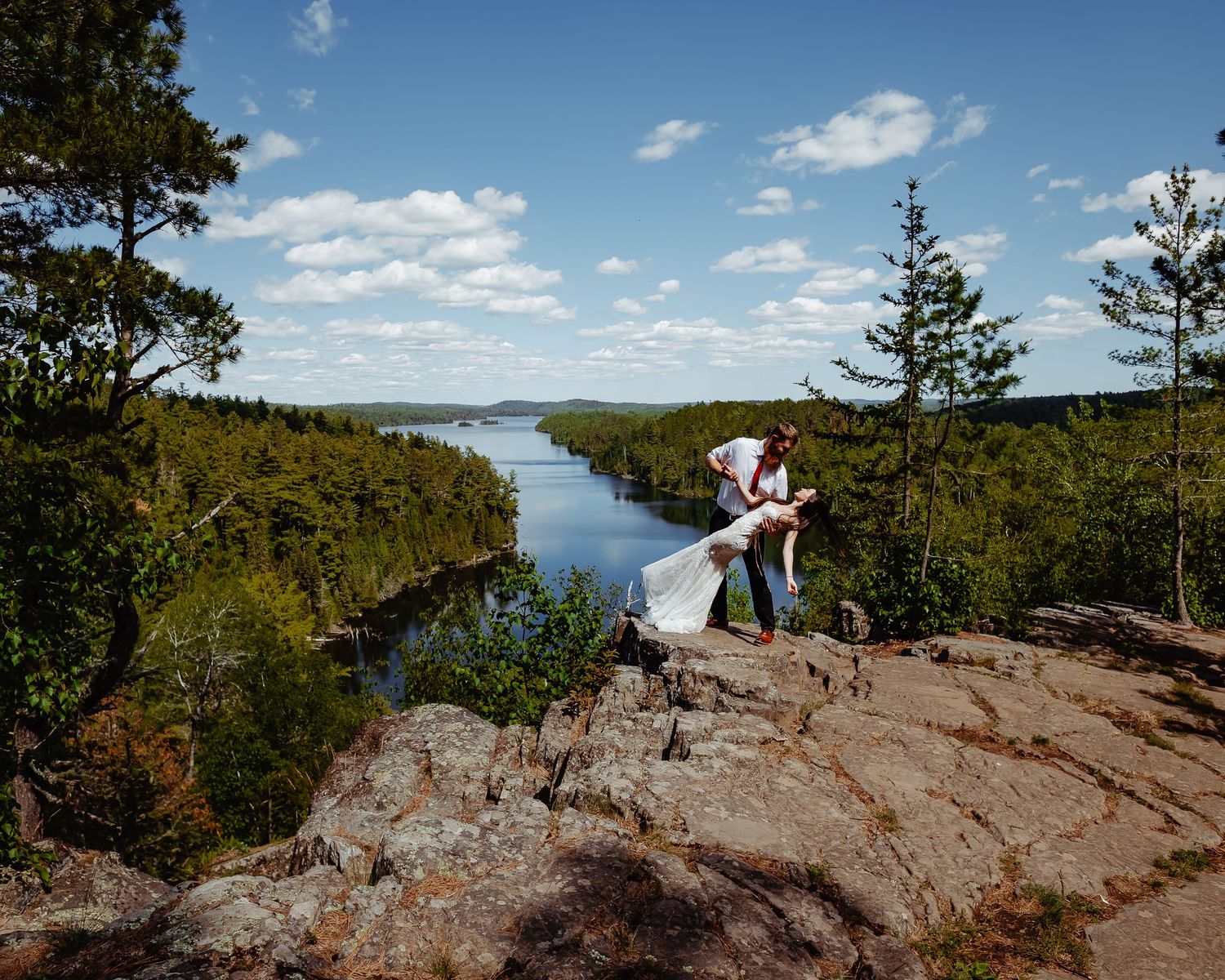 A couple embraces on a rocky cliff overlooking a winding river valley with evergreen forests and blue sky.