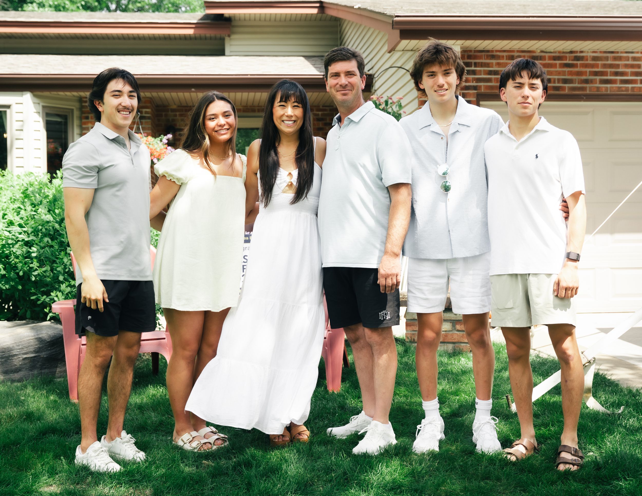 Group of friends dressed in white and gray summer outfits pose together on a lawn in front of a house.
