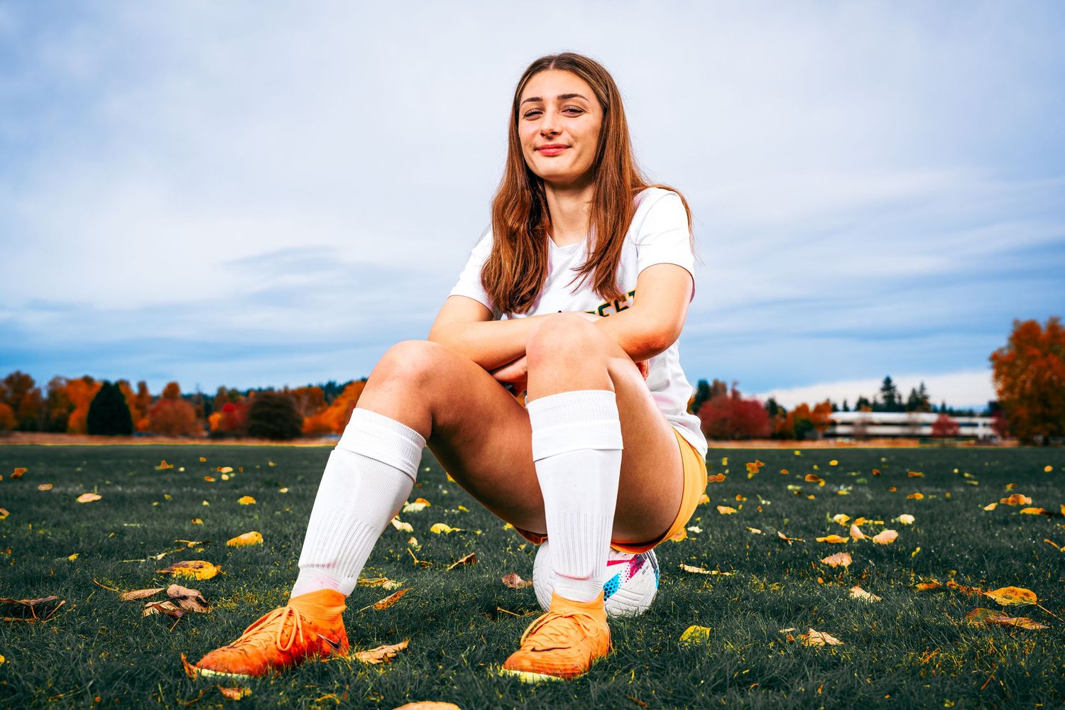 A female soccer player in the Seattle, Washington area sits on a ball in a grassy field covered with autumn leaves.