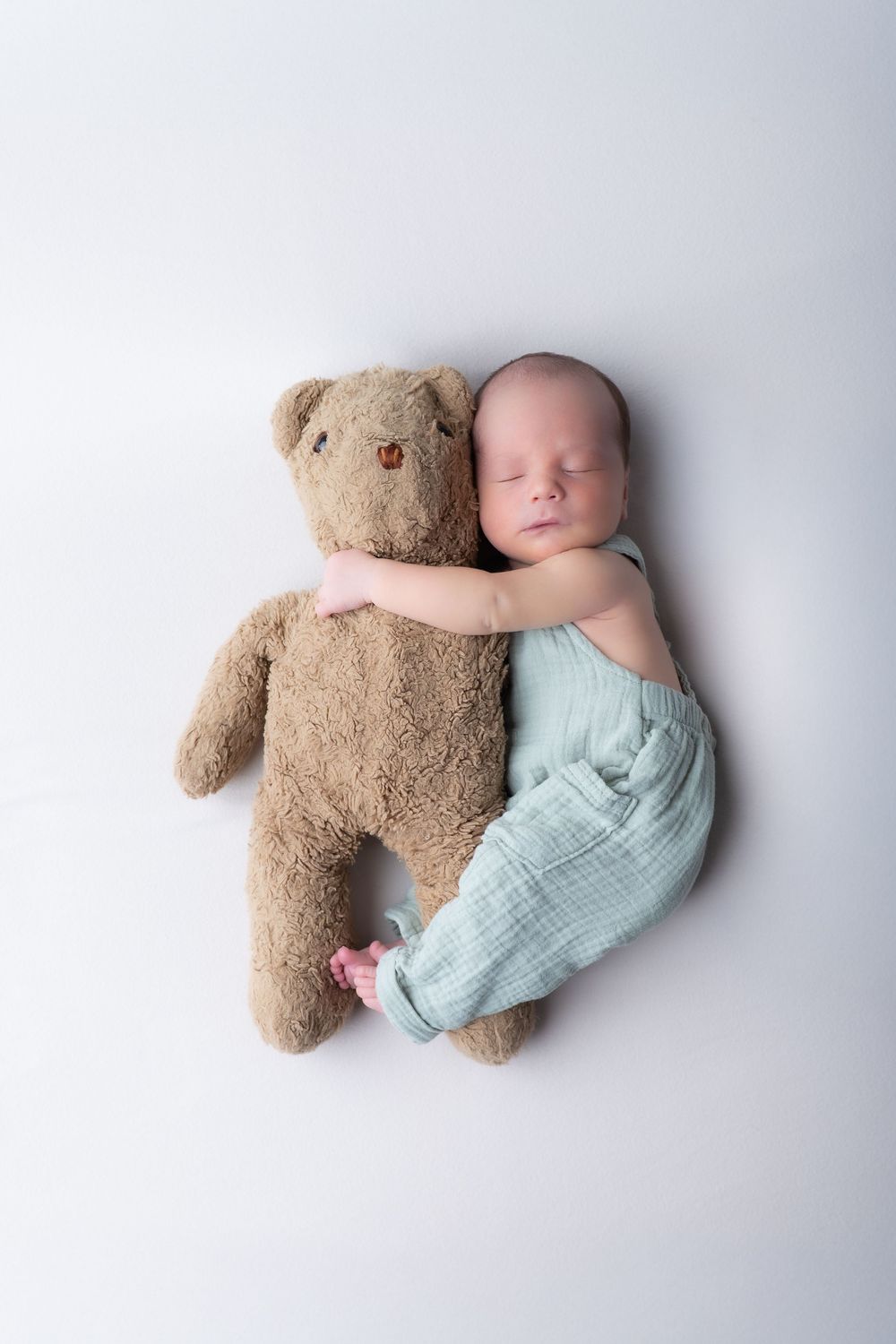 Sleeping newborn baby in mint romper cuddling a brown teddy bear on white background.