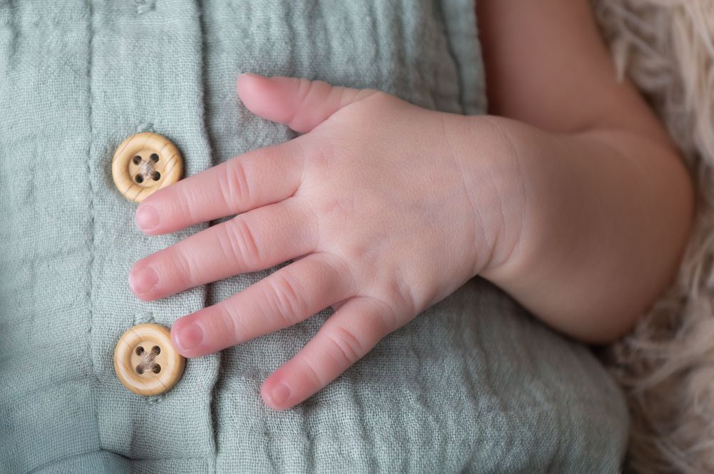 Close-up of a newborn baby's tiny hand resting on a light blue textured romper with wooden buttons.