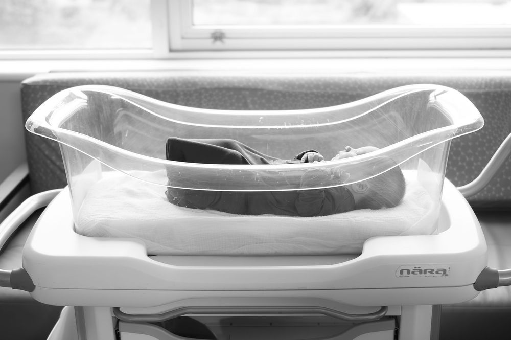 Black and white photo of an empty Nuna baby bassinet on a stand near a window.