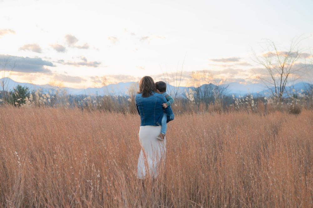 A mother holds her baby while standing in a golden field at sunset with mountains in the background.