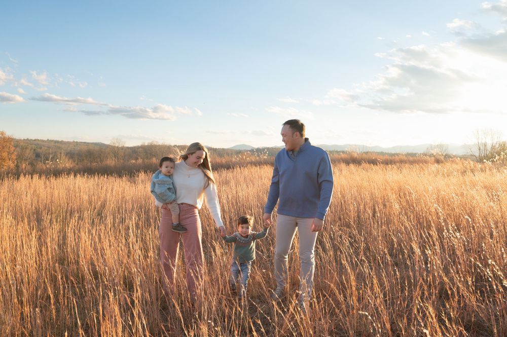 Family of four walking through golden wheat field at sunset, parents holding hands with young children under blue sky.
