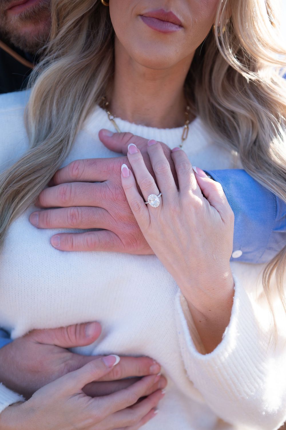 Close-up of a woman's hand displaying a large oval diamond halo engagement ring while embracing her partner outdoors.