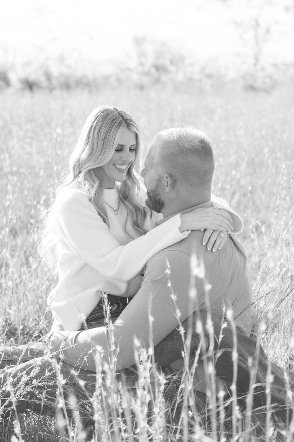 Black and white photo of a smiling couple sitting in a field of tall grass, sharing a romantic moment.