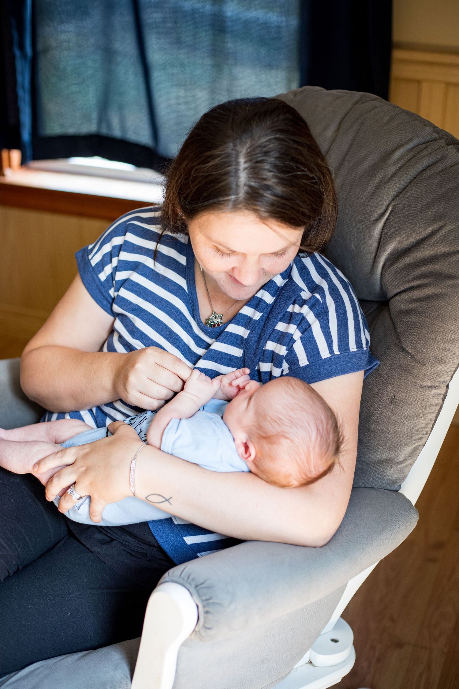 A mother in a striped shirt sits in a gray armchair lovingly gazing at her newborn baby.