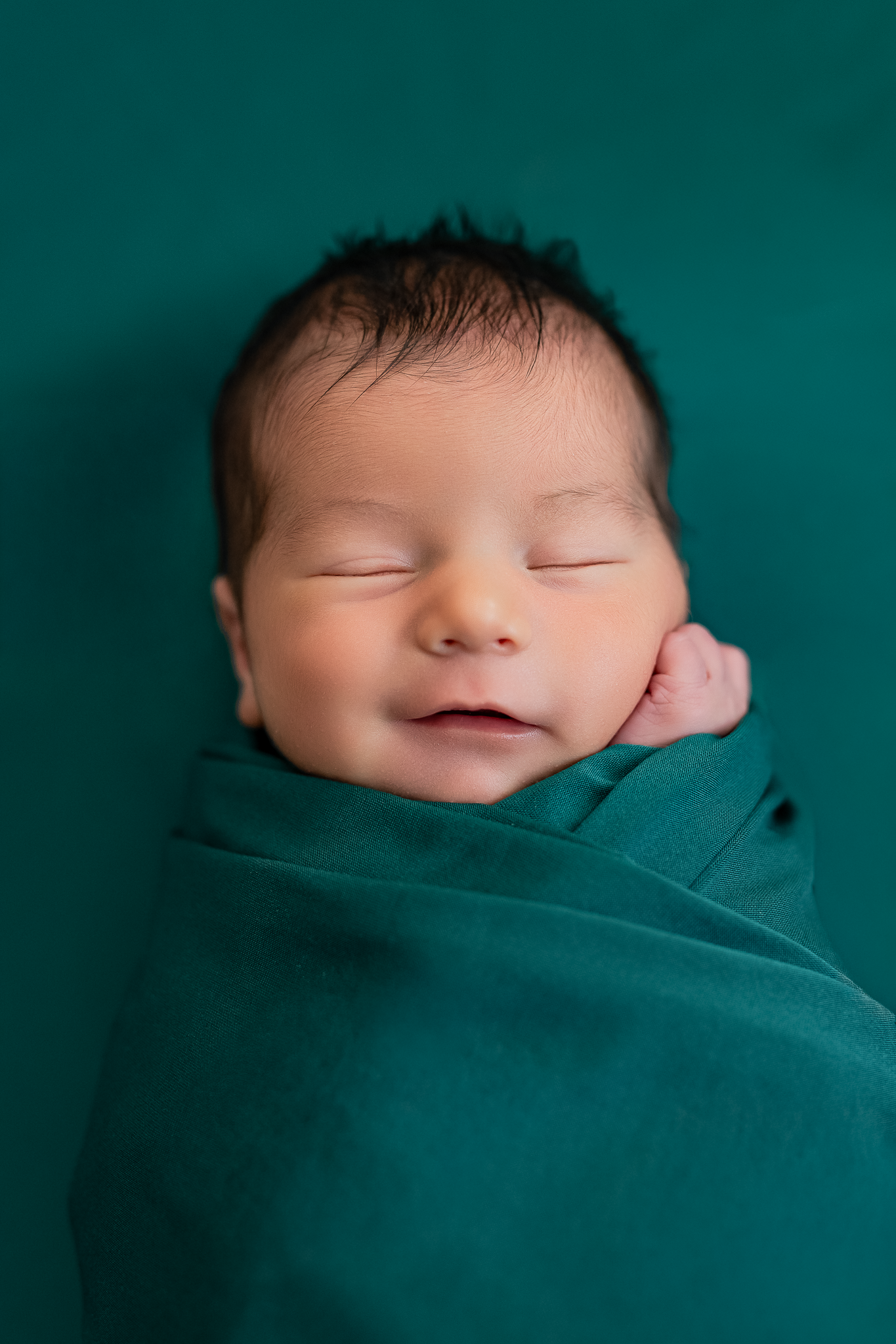 Peaceful newborn baby wrapped in teal swaddle sleeping on soft gray blanket.
