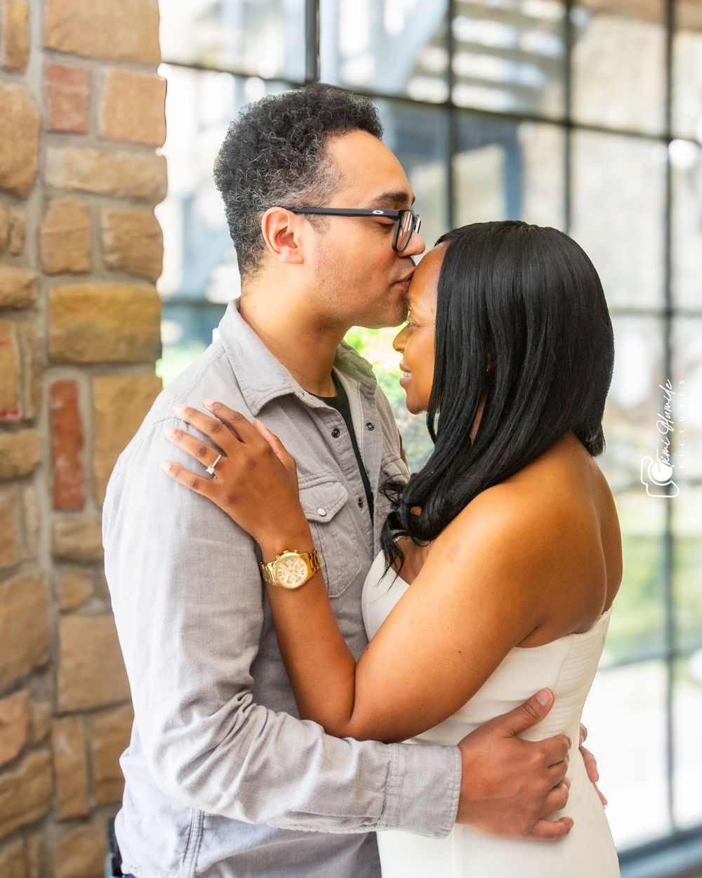 A romantic engagement photo of a couple embracing against a brick wall backdrop.