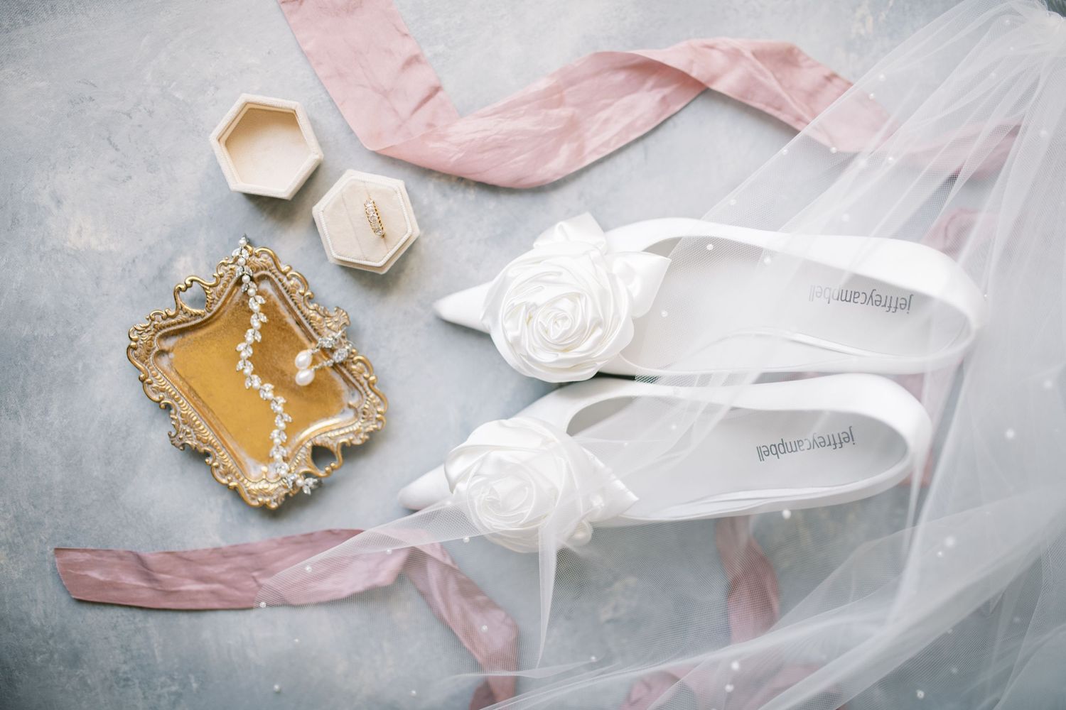 Elegant wedding accessories including white bridal sandals, blush ribbon, a gold jewelry box and veil on light gray surface.
