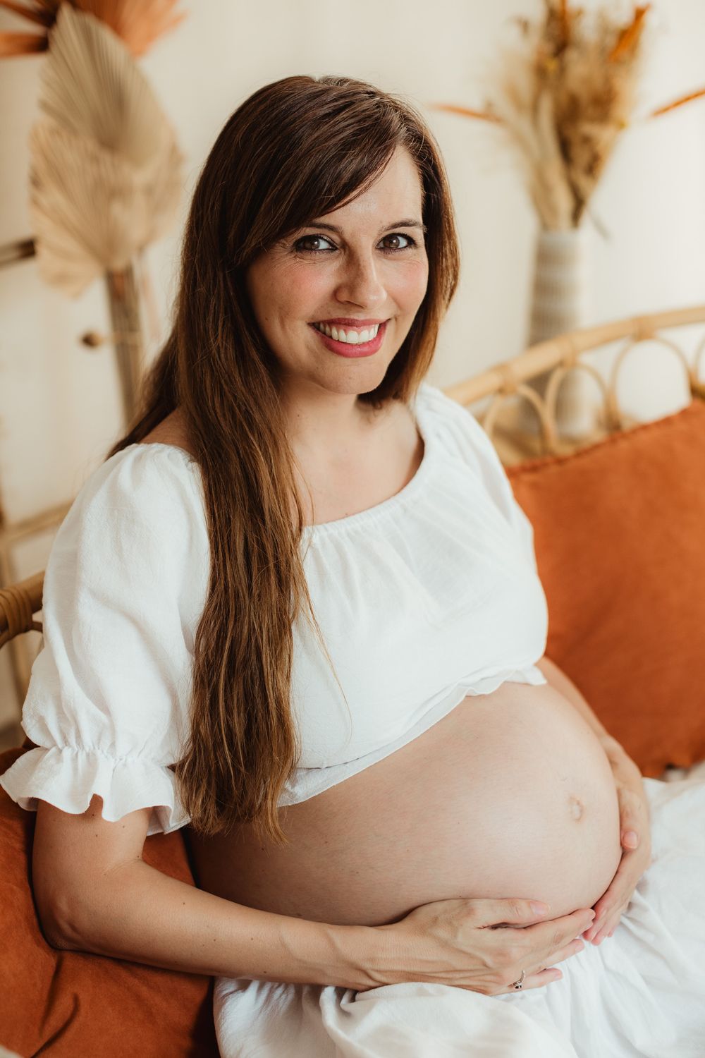 Maternity photo series showing a pregnant woman in white dress against warm neutral backdrop with dried pampas grass decor.