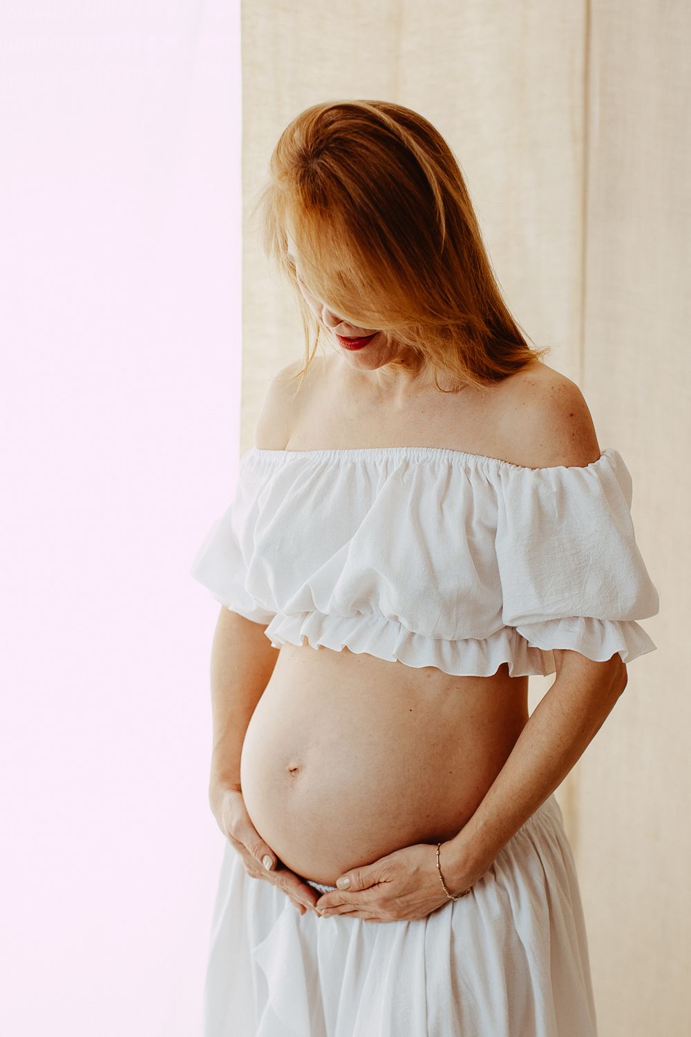 A pregnant woman in a white off-shoulder dress holds her belly while standing against a light background.
