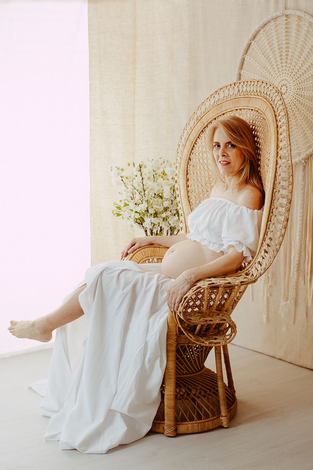 A person in a flowing white dress reclines in a woven peacock chair against a light backdrop.