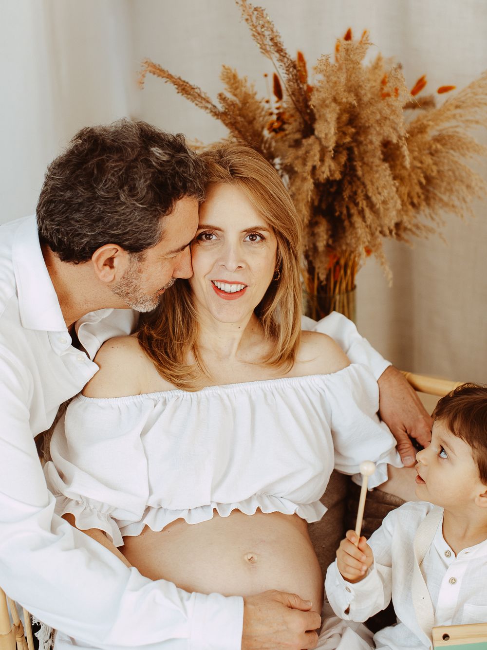 Romantic couple in white clothing embraces tenderly against neutral backdrop with dried pampas grass decor.