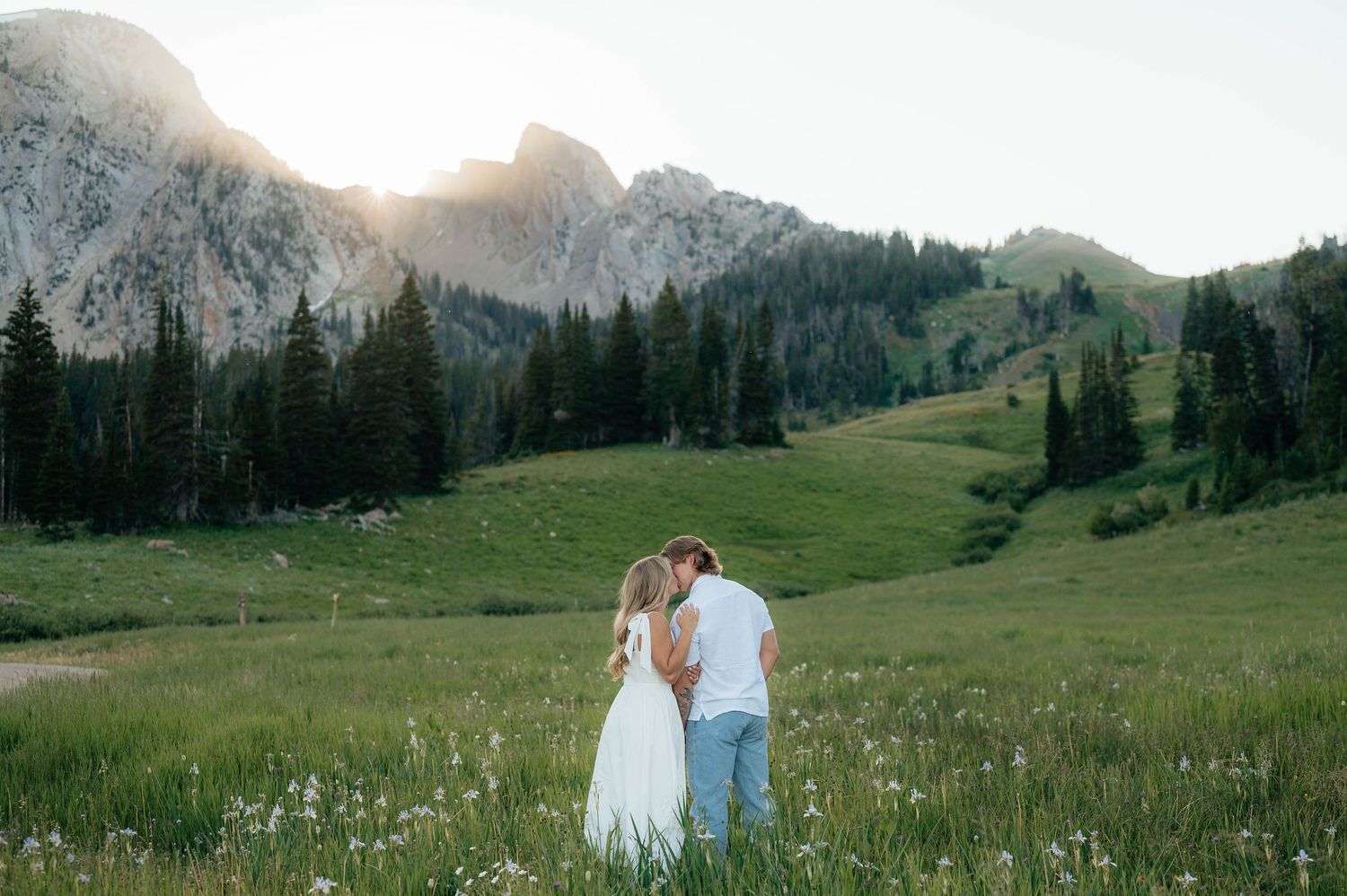 Couple sharing a tender moment in a sunlit wildflower meadow surrounded by mountains and pines.