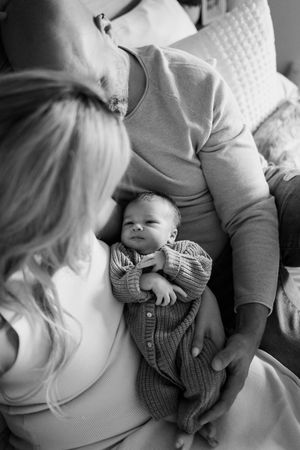 A tender black and white photo of a newborn baby in a striped outfit lying between two adults.