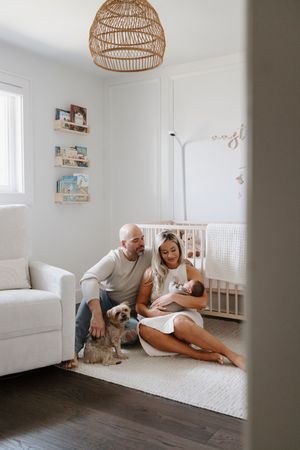 Bright nursery room with rattan lamp, white crib, and family relaxing on floor with dog.