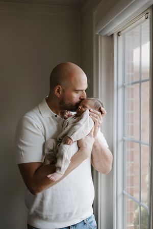 Tender moment by a window as an adult cradles a newborn baby wrapped in white.