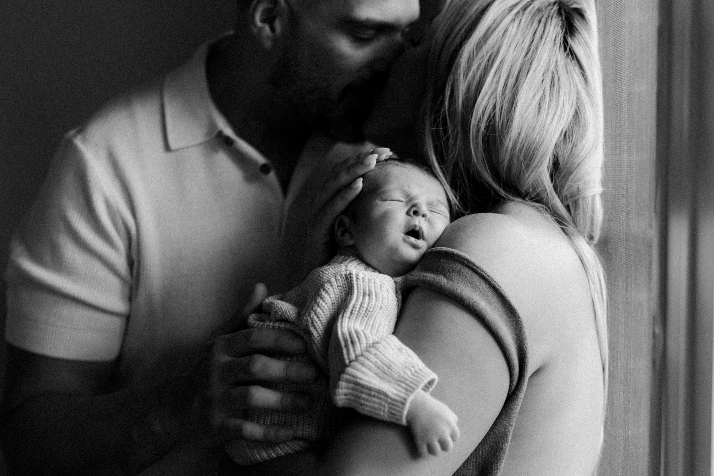 Black and white image of a sleeping newborn being tenderly held by two adults near a window.