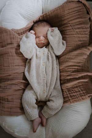 Sleeping newborn wearing a cream knit onesie lying on brown bedding.