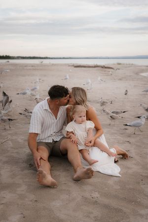 Family relaxes together on sandy beach with seagulls nearby.
