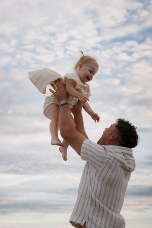 Parent lifts child in white clothing against cloudy sky.
