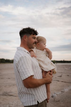 Parent holds child in striped shirt on beach during sunset.