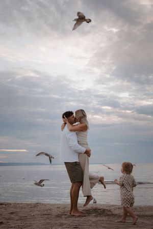 Family plays with seagulls on beach during cloudy sunset.