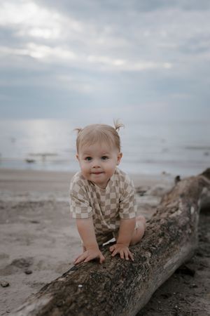 A toddler in checkered clothing plays on a driftwood log at the beach.