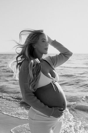 Black and white maternity portraits by ocean waves with wind-swept hair.