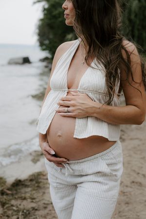 Close-up shots of a pregnant belly in a white ribbed dress on a beach.