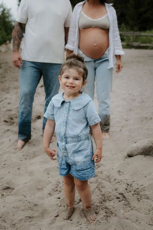 Small child wearing denim outfit standing barefoot in sand at beach.