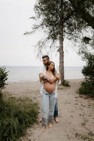 Couple embracing on sandy beach with pine trees in background.