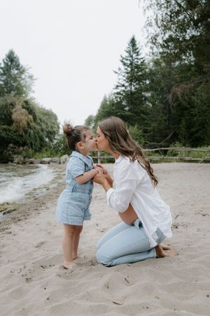 Sweet family moment captured on sandy beach with evergreen trees in background.