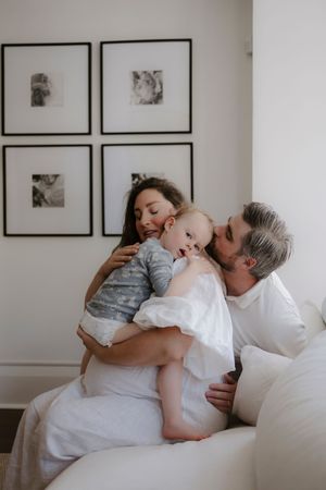 A tender moment shared between parent and child lying on a white bed with framed artwork on the wall.