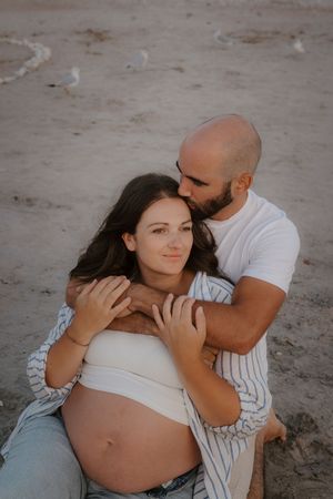 Romantic maternity photos of couple embracing on beach in casual attire.