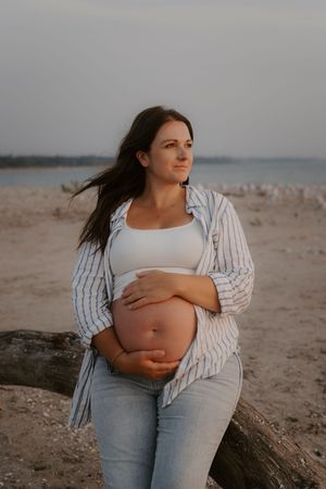 Maternity photos on a beach with woman in white top and striped shirt showing her pregnant belly.