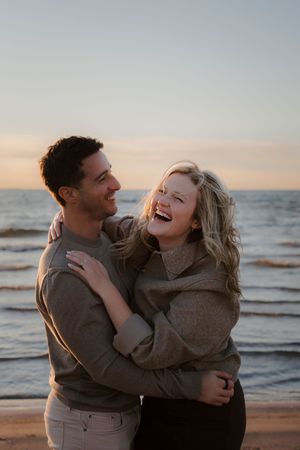 Joyful couple laughing and embracing on beach at sunset wearing matching sweaters.