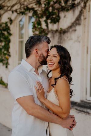 Romantic couple sharing intimate moments against a light colored wall with greenery in an outdoor setting.