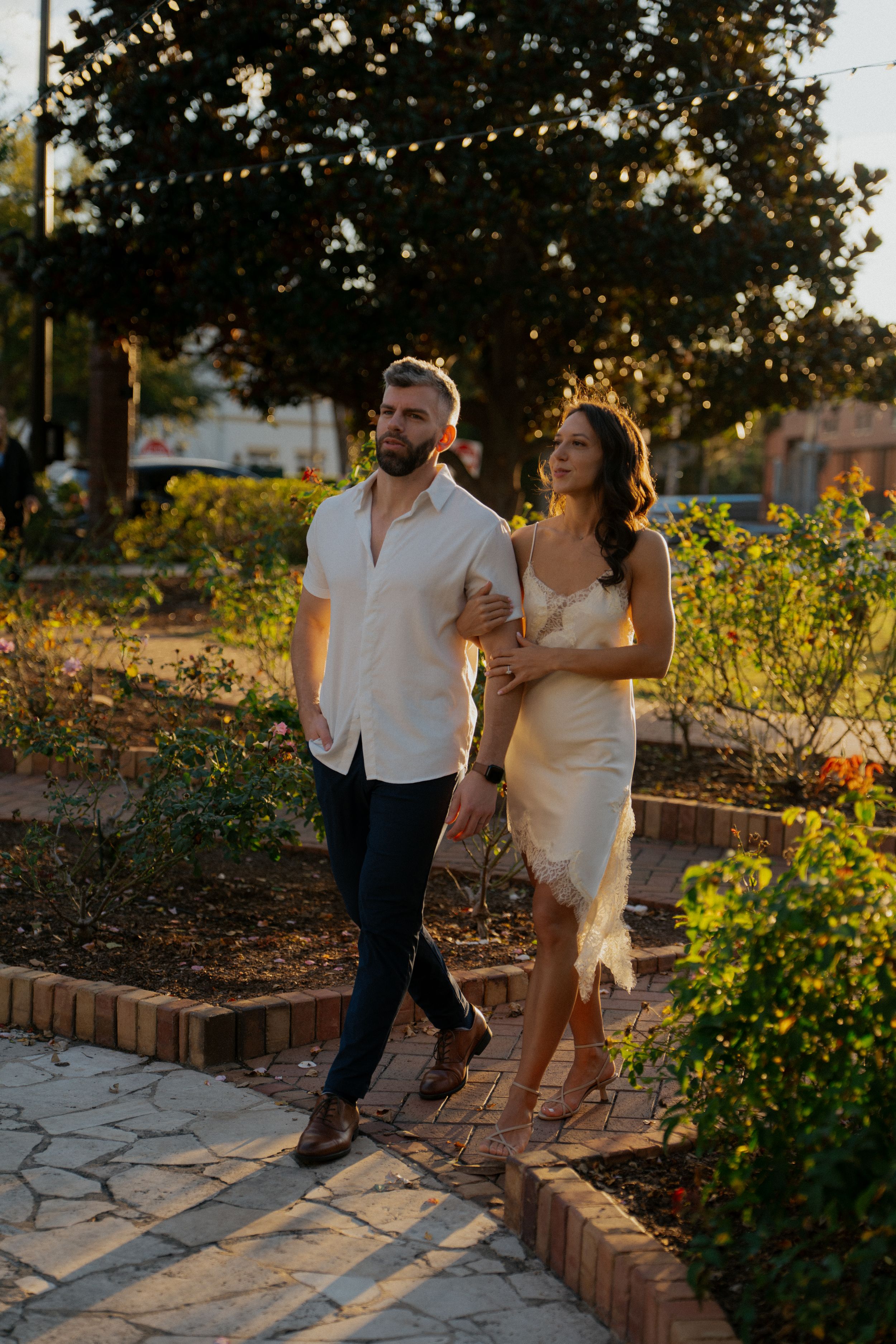 A couple strolls together through a garden path at sunset with warm golden lighting.