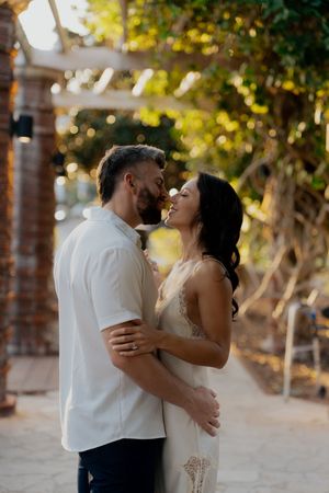Couple in white attire sharing tender moments outdoors with sunlit trees in the background.