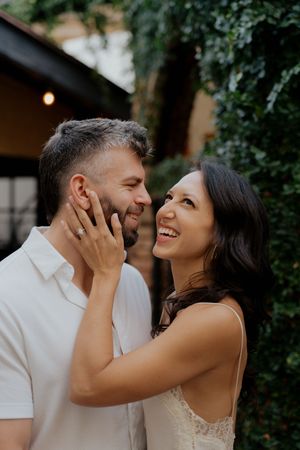 A couple shares joyful laughter and tender touches during an outdoor photoshoot with greenery in the background.