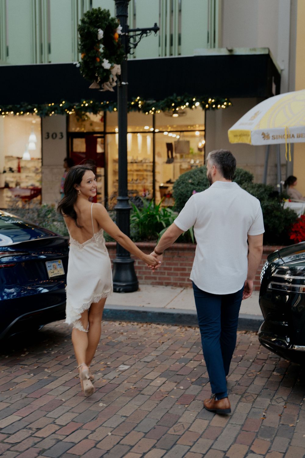 A couple holds hands while walking down a charming brick-paved street with storefronts.