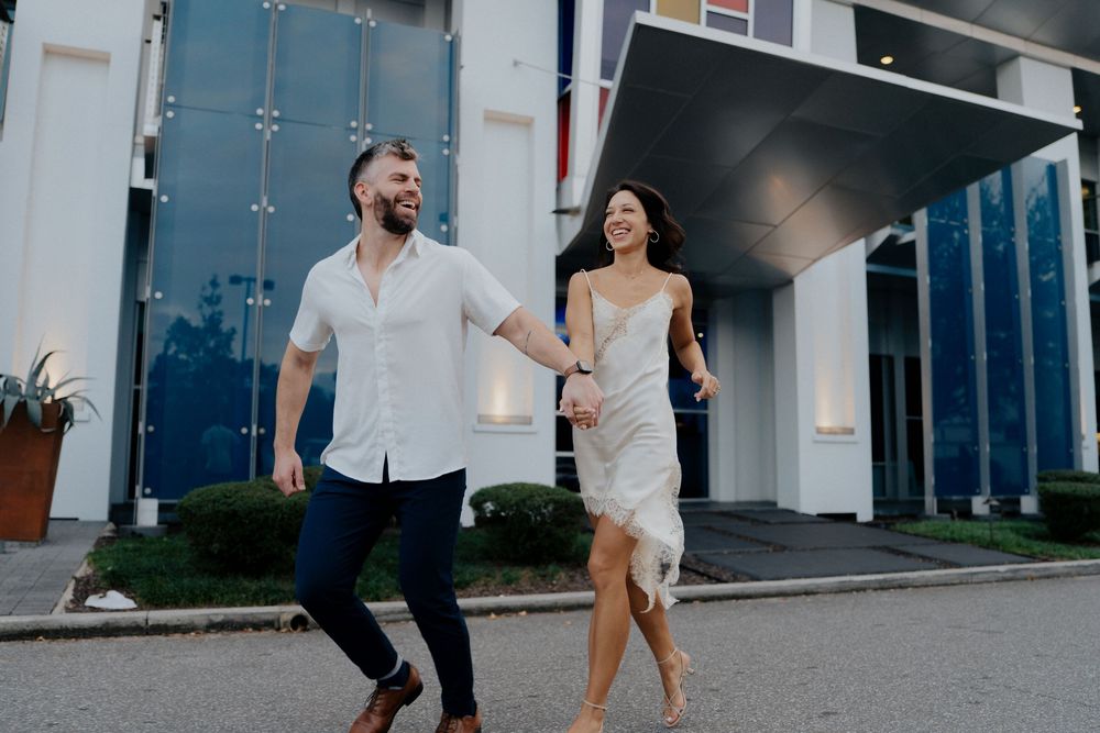 A couple in casual attire dances together on a sidewalk outside a modern building.