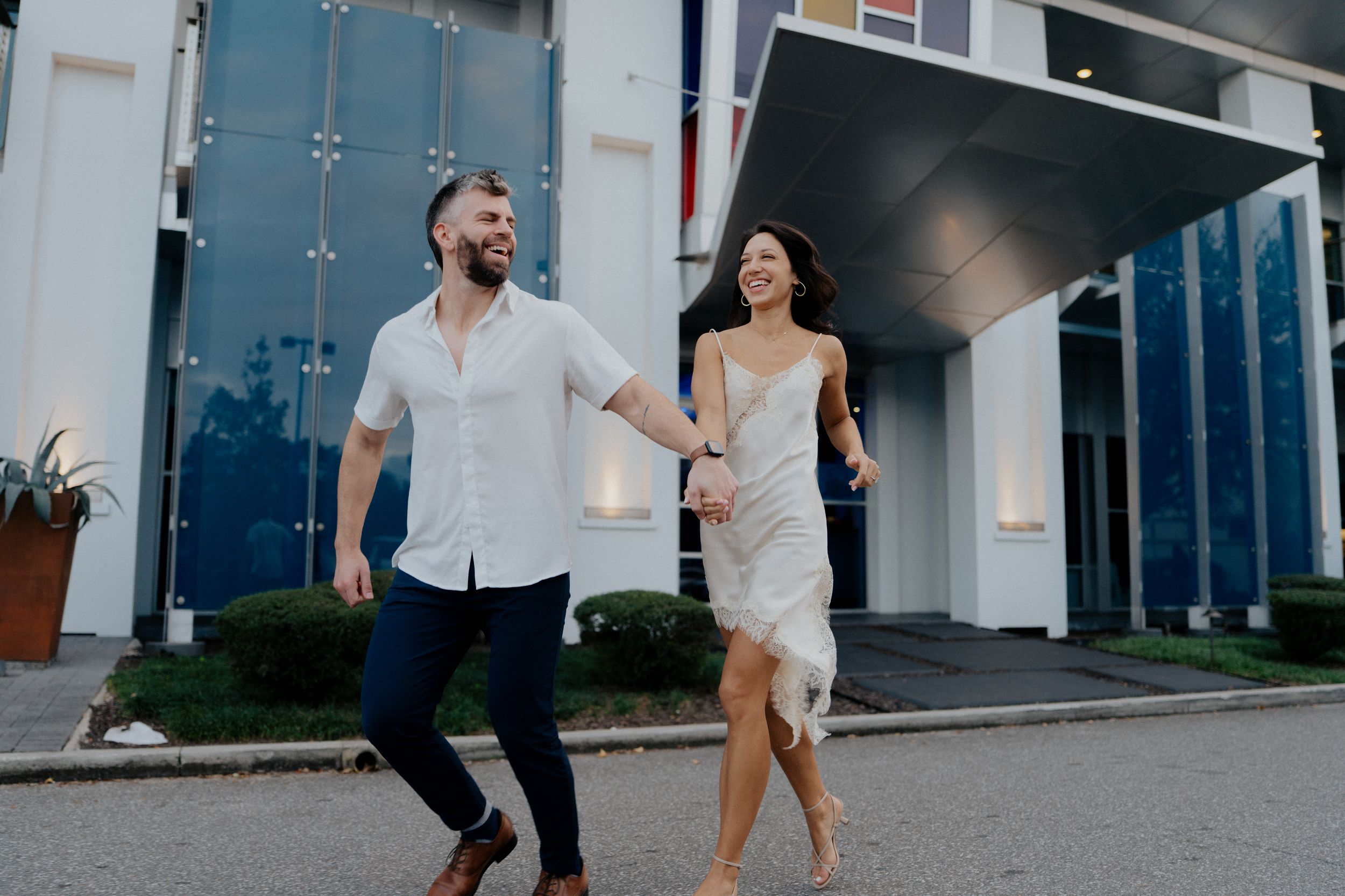 A couple in casual attire dances together on a sidewalk outside a modern building.
