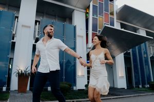 Couple in white outfits dances together during a romantic photoshoot session outside a modern building.