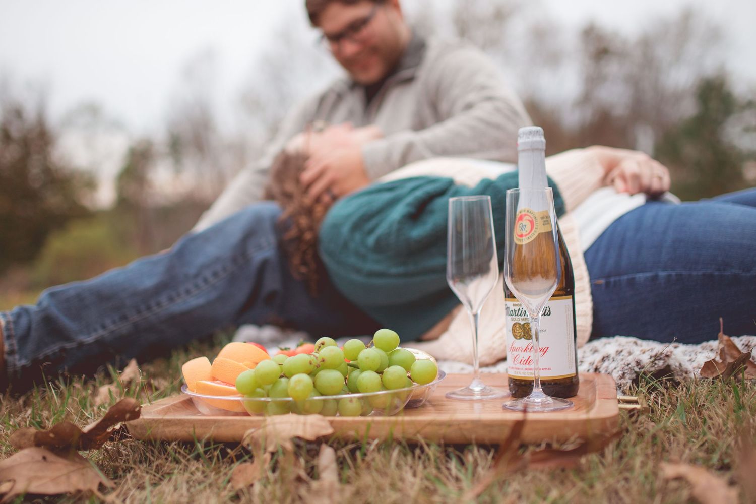 Romantic picnic scene with wine, fresh fruits and couple relaxing in background.