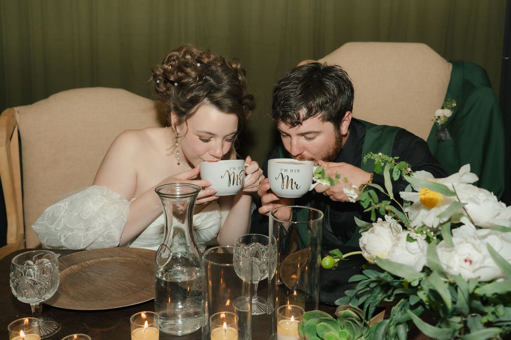 Bride and groom sipping from Mr. and Mrs. cups at candlelit wedding reception table with white floral arrangements.